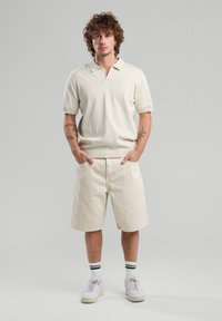Beige short-sleeve polo shirt with a small collar, paired with knee-length beige shorts. White sneakers and green-striped socks.