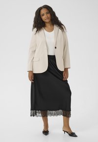 Vaaleanbeige blazer, jossa on viistot läpät, valkoisen paidan päällä, mustan satiinihameen parissa, jossa on pitsireuna, sekä mustat kärkitossut.