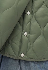 Jachetă verde cu umplutură, având nasturi argintii plați, o suprafață netedă și un model texturat în material. Margină cu o cusătură contrastantă.