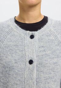 Világosszürke kötött kardigán, mintás textúrával, kerek nyakkivágással és három fekete gombbal elöl. Puha, bolyhos anyagból.