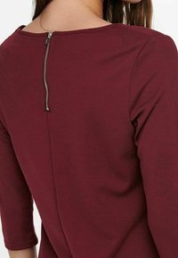 Haut bordeaux avec un col rond, des manches trois-quarts et une fermeture éclair au dos avec des détails en métal. Confectionné dans un tissu lisse.