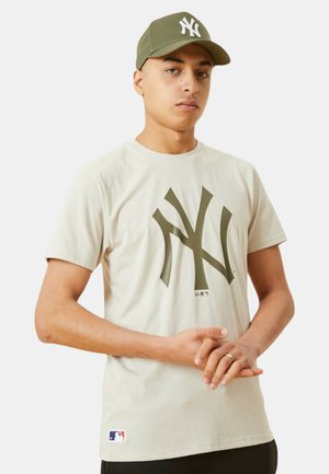 T-shirt in cotone beige con un grande logo NY verde scuro sul davanti. Abbinato a un cappellino da baseball verde coordinato, entrambi pezzi di abbigliamento casual.