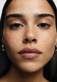 Närbild av ett ansikte med ljusbrun hy, synliga fräknar, fylliga läppar med en nude-gloss, och mörkt hår. Smycken är silverörhängen i form av hooper.