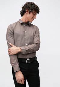 Braunes Hemd mit durchgehender Knopfleiste, langen Ärmeln, subtiler Textur und weißen Knöpfen. Getragen mit schwarzen Hosen und einem Gürtel, zeigt es ein tailliertes Design.