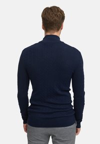 Granatowy sweter w splot warkoczowy z długimi rękawami, ściągaczami przy mankietach i u dołu, z teksturowanym wzorem. Noszony z szarymi spodniami.