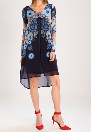 Femme portant une robe longue à manches longues fleurie bleu marine avec un ourlet transparent et des chaussures à talons hauts rouges vives à bride à la cheville, debout devant un fond uni.