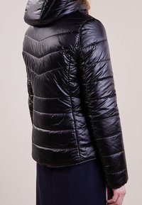 Veste en duvet noire avec capuche, design matelassé en chevrons et finition brillante. Coupe structurée avec des poignets et un ourlet élastiques.
