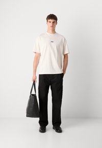 Helle beigefarbene Baumwoll-T-Shirt mit kleinem schwarzem Logo, kombiniert mit weit geschnittenen schwarzen Cargo-Hosen und einer schwarzen Ledertasche.