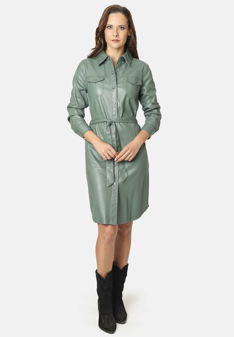 Vestido de camisa de piel sintética verde con mangas largas, cierre de botones en la parte delantera, dos bolsillos en el pecho y cinturón a juego. Combinado con botas de vaquero negras.