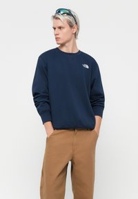 Navy blue sweatshirt with long sleeves and ribbed cuffs, featuring a small white logo on the chest. Tan pants are worn below.