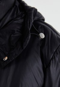 Schwarze Pufferjacke mit gestepptem Design, Kapuze mit verstellbarem Kordelzug und metallischen Druckknopfdetails am Kragen.
