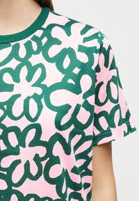 T-skjorte med grønt abstrakt blomstermønster på lyserosa bakgrunn, båret av en person som viser overkroppen og kort erme.