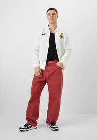Hvid zip-jakke med gule detaljer og logo, sort t-shirt, røde løse jeans, sort og hvidt sneakers. Enkel baggrund.