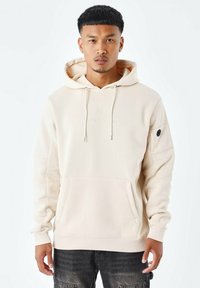 Sudadera con capucha beige que presenta un bolsillo frontal, cordones, acentos laterales y un sutil logotipo en el pecho. Fabricada con tejido suave.