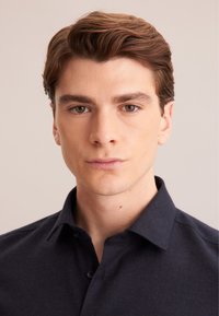 Camicia button-up blu scuro con colletto, realizzata in tessuto testurizzato. Il modello ha capelli castani corti e ben pettinati, con un'espressione neutra.