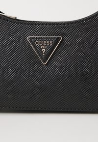 Sac à main noir texturé avec une étiquette de logo triangulaire et des accents dorés. Le matériau semble synthétique avec un motif légèrement en relief.
