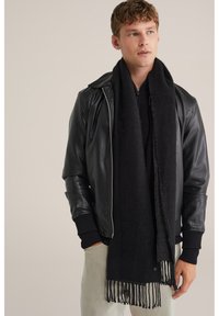 Melnā šalle, izgatavota no mīksta auduma, ar frendžiem beigās. Valkāta virs melnā ādas jakas ar rāvējslēdzēju un tumšā adītā džempera.