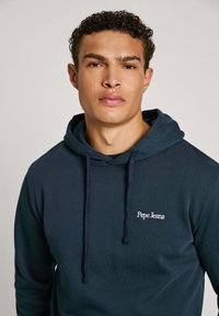 Felpa blu navy in tessuto morbido, con tasca frontale, cappuccio con cordino e logo ricamato bianco "Pepe Jeans" sul petto.
