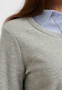 Maglione grigio chiaro e morbido con collo rotondo; indossato sopra una camicia a righe blu e bianche con collo. Texture liscia, design casual.
