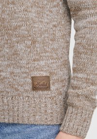 Strikket sweater i brune og creme farver med ribbet kant og manchetter, samt et lædermærke med teksten "Solid" foran.