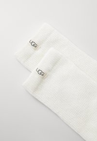 Hvite strikkede leggmuffer med UGG-merkelapp; strukturert mønster; sylindrisk form; mykt materiale.