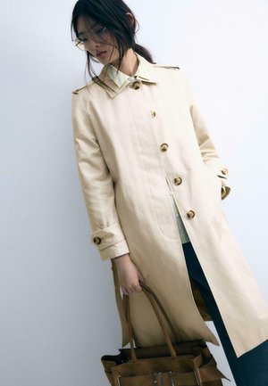 Kvinde iført beige trenchcoat, der holder en brun lædertaske, iført briller og en t-shirt med striber, stående foran en ensartet hvid baggrund.