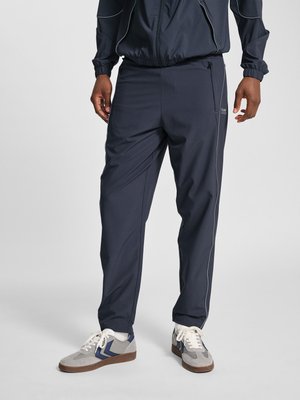 Osoba w czarnych spodniach dresowych i szarych butach sportowych z niebieskimi akcentami, stojąca na tle jednolitego jasnego tła.