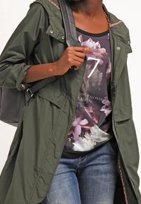 Persona vestida con una chaqueta verde con capucha sobre una camisa de estampado floral y jeans azules, llevando un bolso negro de hombro.