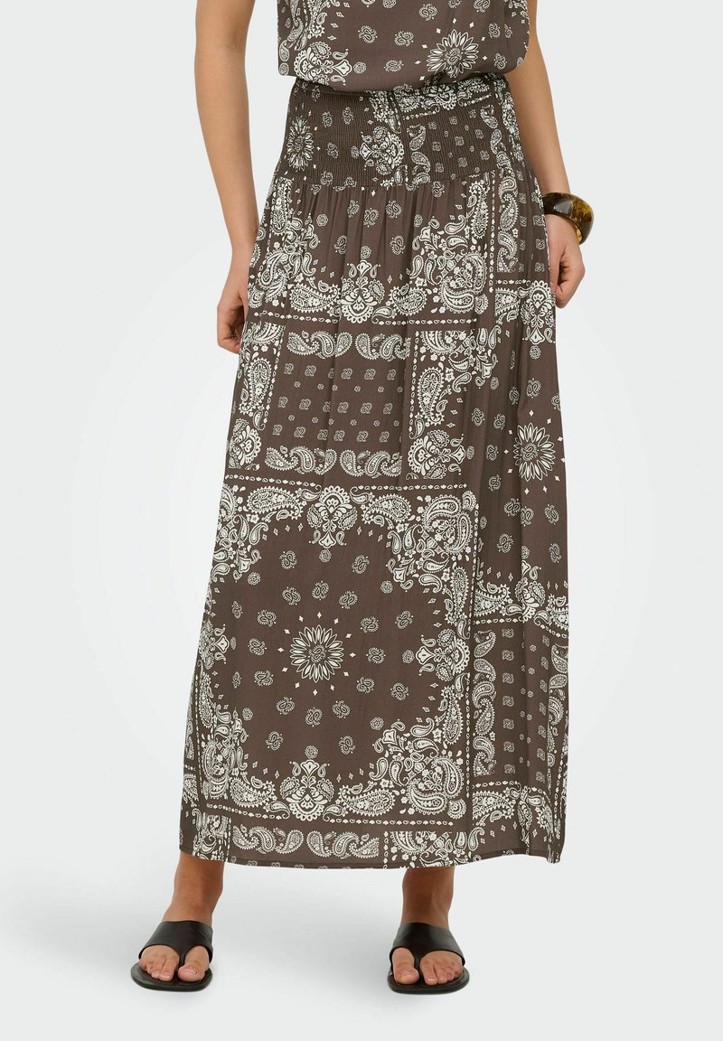 Femme portant une jupe maxi marron avec un imprimé bandana blanc paisley et floral, associée à des sandales noires à bout ouvert et un bracelet marron.