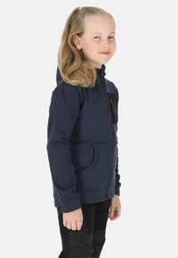 Marineblauwe hoodie met ritssluiting, kangoeroezak, gestructureerde stof en capuchon. Zwarte accenten op de borst. Draagt zwarte broek.