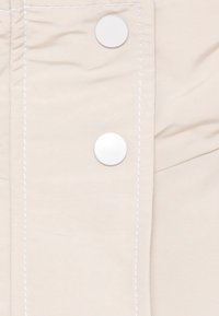 Tela beige con una textura suave que cuenta con tres botones de presión blancos y costuras en blanco contrastantes a lo largo de las costuras.
