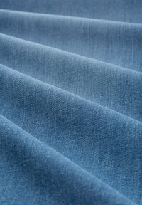 Svijetloplavi denim materijal s glatkom teksturom, koji prikazuje suptilne promjene boje i dijagonalni uzorak tkanja. Materijal je presavijen, što stvara dubinu.