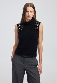 Czarny bezrękawnik z golfem z puszystego materiału, zdobiony kryształowymi aplikacjami na ramionach, zestawiony z szarymi spodniami.