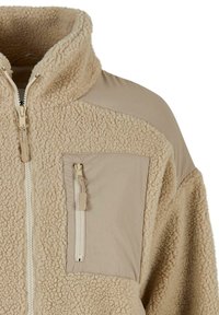 Bež flis jakna s visokim ovratnikom, prednjim patentnim zatvaračem i džepom na prsima s patentom, s glatkim platnenim panelima na ramenu i džepu.