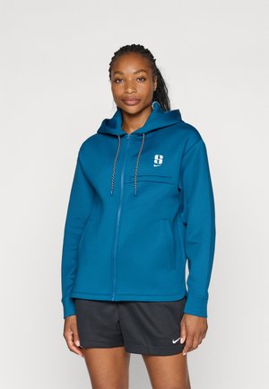 Teal hettegenser med glidelås, snøring på hetten, frontlomme og en liten logo på brystet. Myk tekstur, kombinert med svarte shorts.