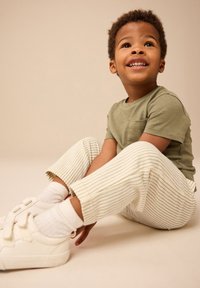 Otrok, oblečen v svetlo zeleno kratkoročno majico in kremno črtaste hlače, obut v bele superge in gležnijske nogavice, sedi na nevtralnem ozadju.