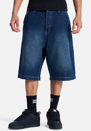 Mężczyzna ubrany w luźne, sięgające kolan ciemnoniebieskie szorty jeansowe, czarne skarpety DC oraz czarne sneakersy DC na białym tle.