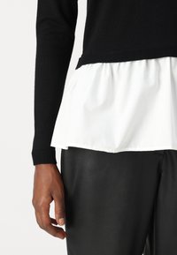 Czarny sweter z ribbowanymi mankietami, nałożony na biały peplum, w połączeniu z dopasowanymi czarnymi spodniami z ekoskóry.