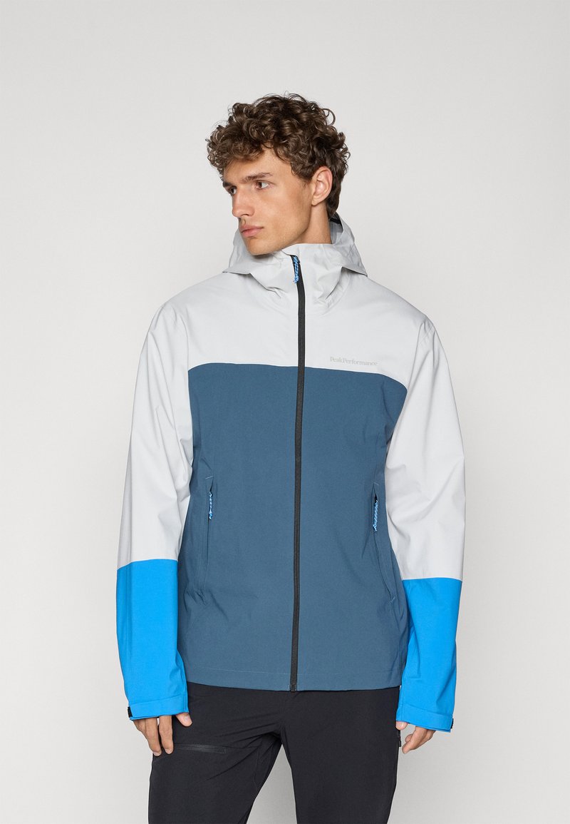 Veste pour homme avec un design en coloris blocs gris, bleu et marine. Matériau imperméable, avec capuche et deux poches latérales zippées.