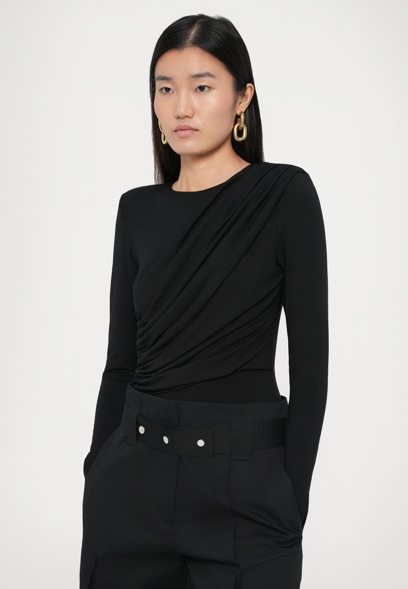 Czarny bodysuit z długim rękawem z drapowanym detalem z przodu. Połączony z czarnymi spodniami o wysokim stanie, wyposażonymi w szeroki pasek i kontrastowe guziki.