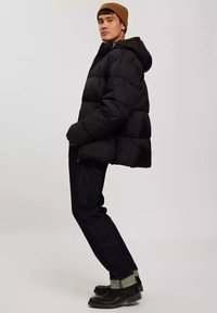 Czarna kurtka puchowa z kapturem, o pikowanym wzorze, z bocznymi kieszeniami, noszona na ciemnych dżinsach i czarnych butach, z brązową czapką. 