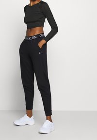 Kobieta w czarnej, dopasowanej bluzie z długim rękawem, czarnych dżoggerach Calvin Klein z logo w pasie i białych sportowych sneakersach, stojąca z rękami w kieszeniach.