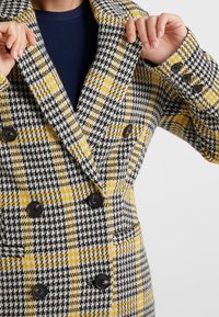 Dvouřadý kabát v černobíložlutém kostkovaném vzoru. Vybaven velkými černými knoflíky a klopami. Měkká, strukturovaná látka.