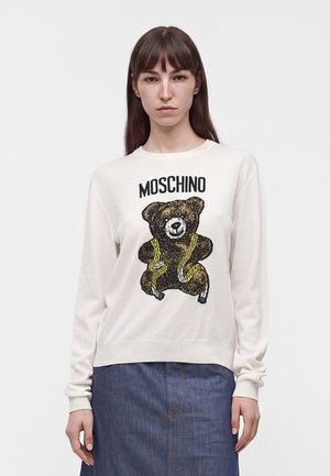 Kvinde med glat brunt hår bærer en hvid Moschino trøje med teddybjørn og målebånd design, kombineret med et blåt denimskørt.