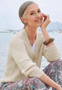 Beige gebreide cardigan met v-hals, knopen en geribde mouwen, gedragen over een witte top, gecombineerd met gestreepte roze en blauwe broeken.