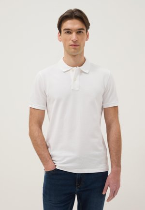 Man draagt een eenvoudige witte korte mouw polo en donkerblauwe spijkerbroek, staat met één hand in de zak tegen een lichte achtergrond.