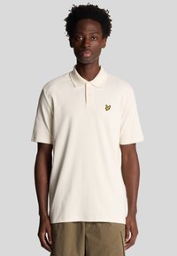 Crème kleurige polo shirt met korte mouwen, een kraag, en een klein geel logo geborduurd op de linkerborst. Structuurstof.