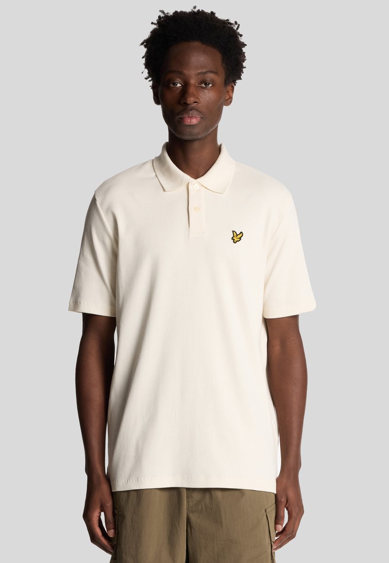 Crème kleurige polo shirt met korte mouwen, een kraag, en een klein geel logo geborduurd op de linkerborst. Structuurstof.