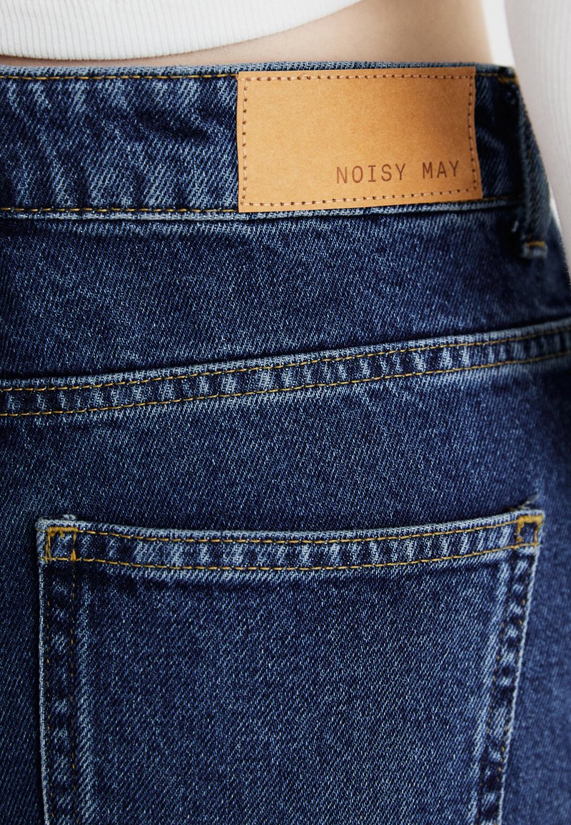 Ciemnoniebieskie dżinsy z denimu mają skórzaną metkę z napisem "NOISY MAY" przy pasku, kontrastowe przeszycia oraz jedną tylną kieszeń.
