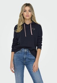 Felpa navy con lacci rosa e ricamo a forma di cuore. Abbinata a jeans blu a vita alta, con una vestibilità rilassata e una trama denim classica.